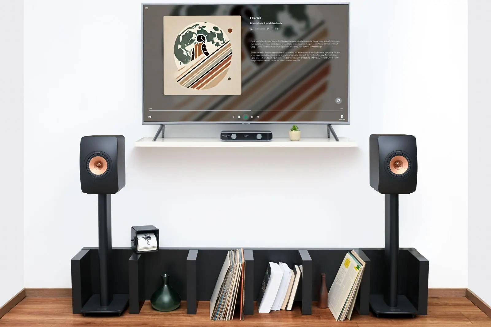 A living room with a TV on the wall showing album art, two speakers on stands and Primo Plus on a low shelf with records.