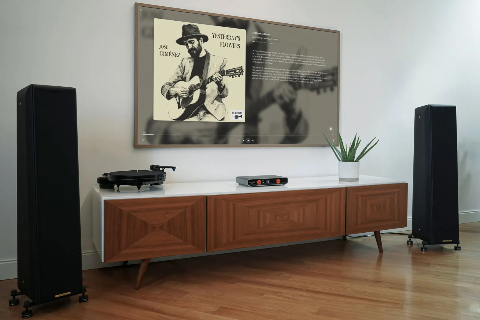A living room with Integro on a wooden sideboard between two tall black floor-standing speakers, next to a turntable and a plant, under a wall TV showing album art.