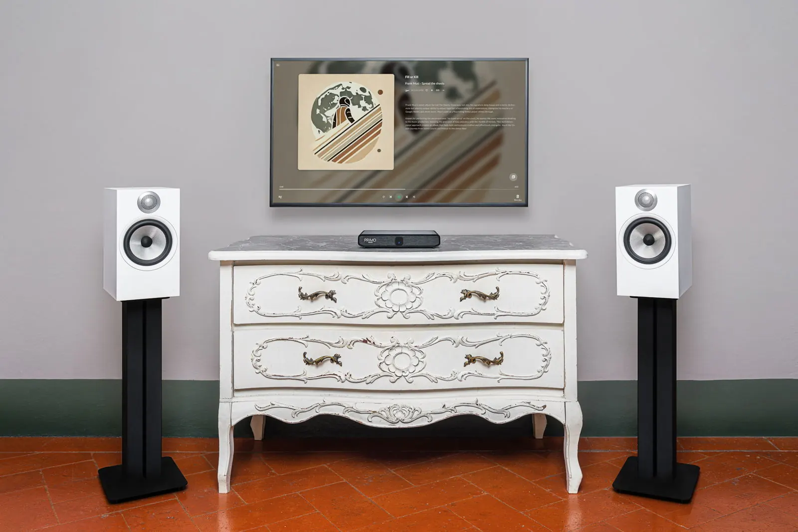 A living room with a TV on the wall showing album art, two white speakers on stands and Primo V3 on a white carved dresser.