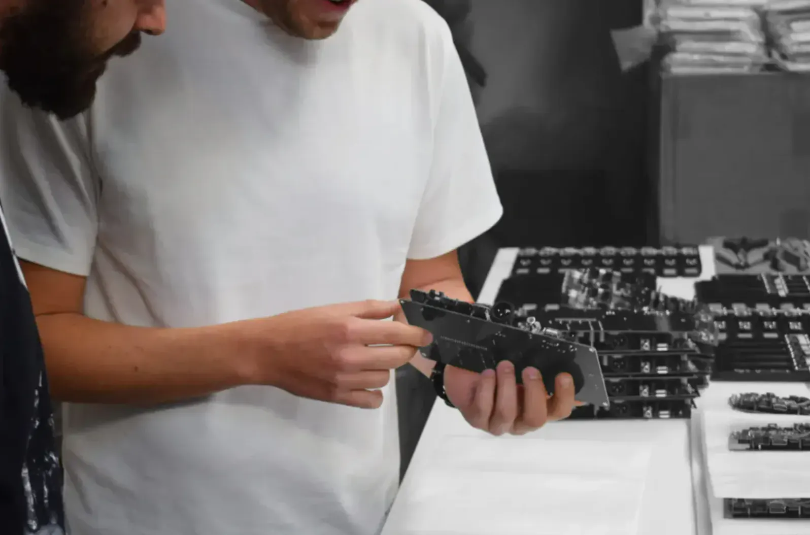 At a workbench, a person in a white T-shirt holds a dark circuit board above rows of more boards.
