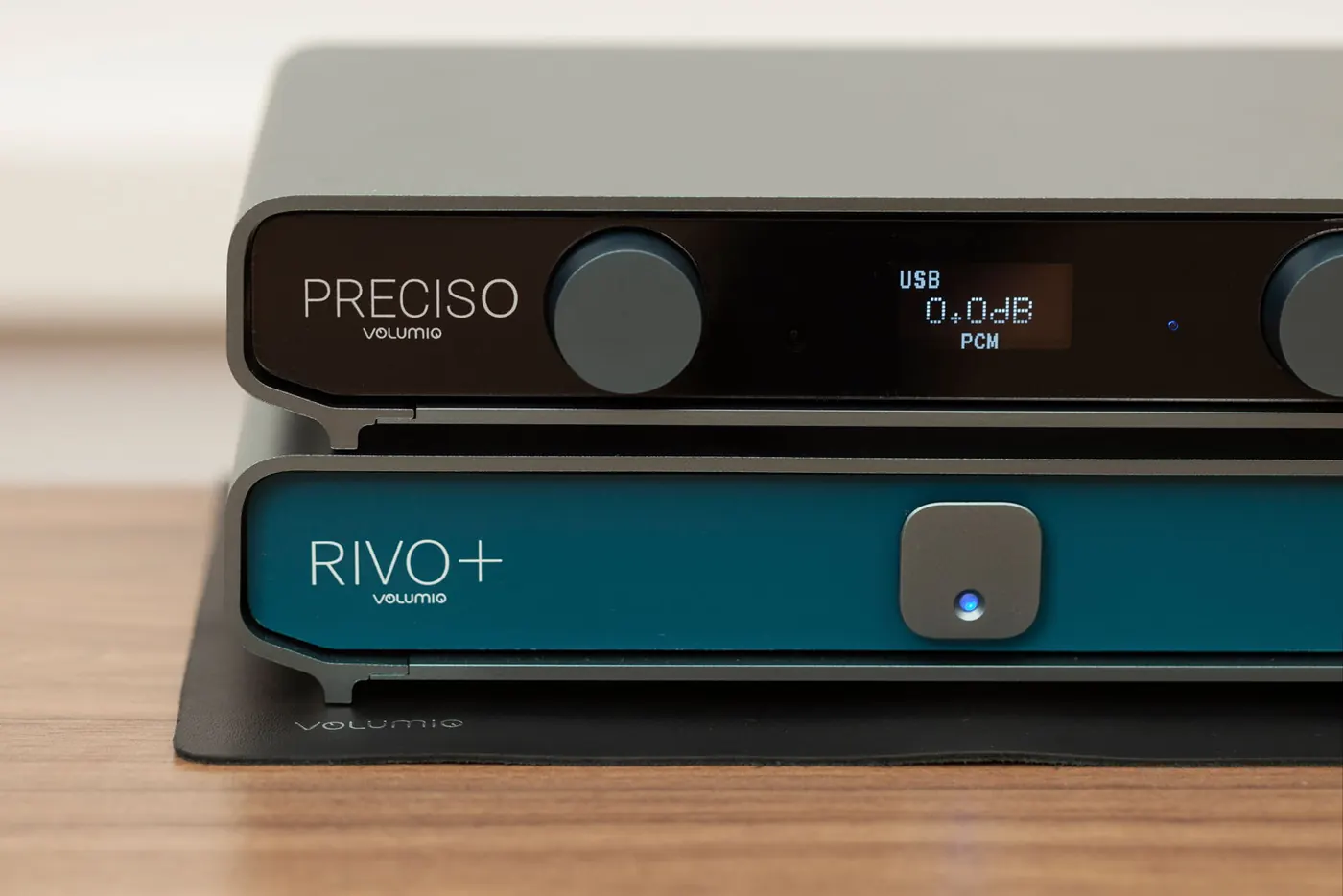 Preciso stacked on top of a Rivo+ with a blue front, on a black mat on a wooden table. The Preciso display reads USB, 0.0dB and PCM.