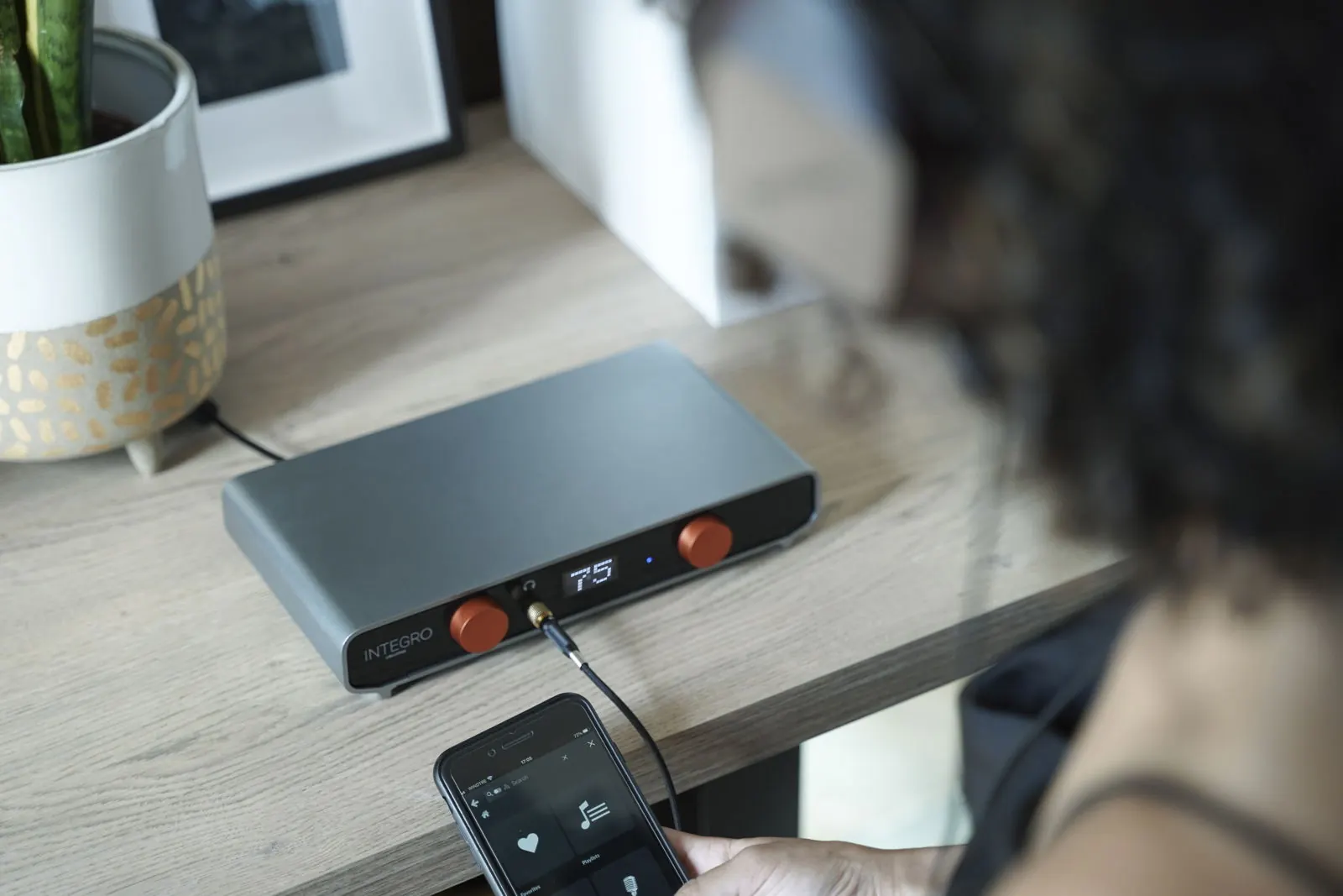A listener holds a phone that shows a music library, next to Integro on a wooden table, with headphones plugged into the front.