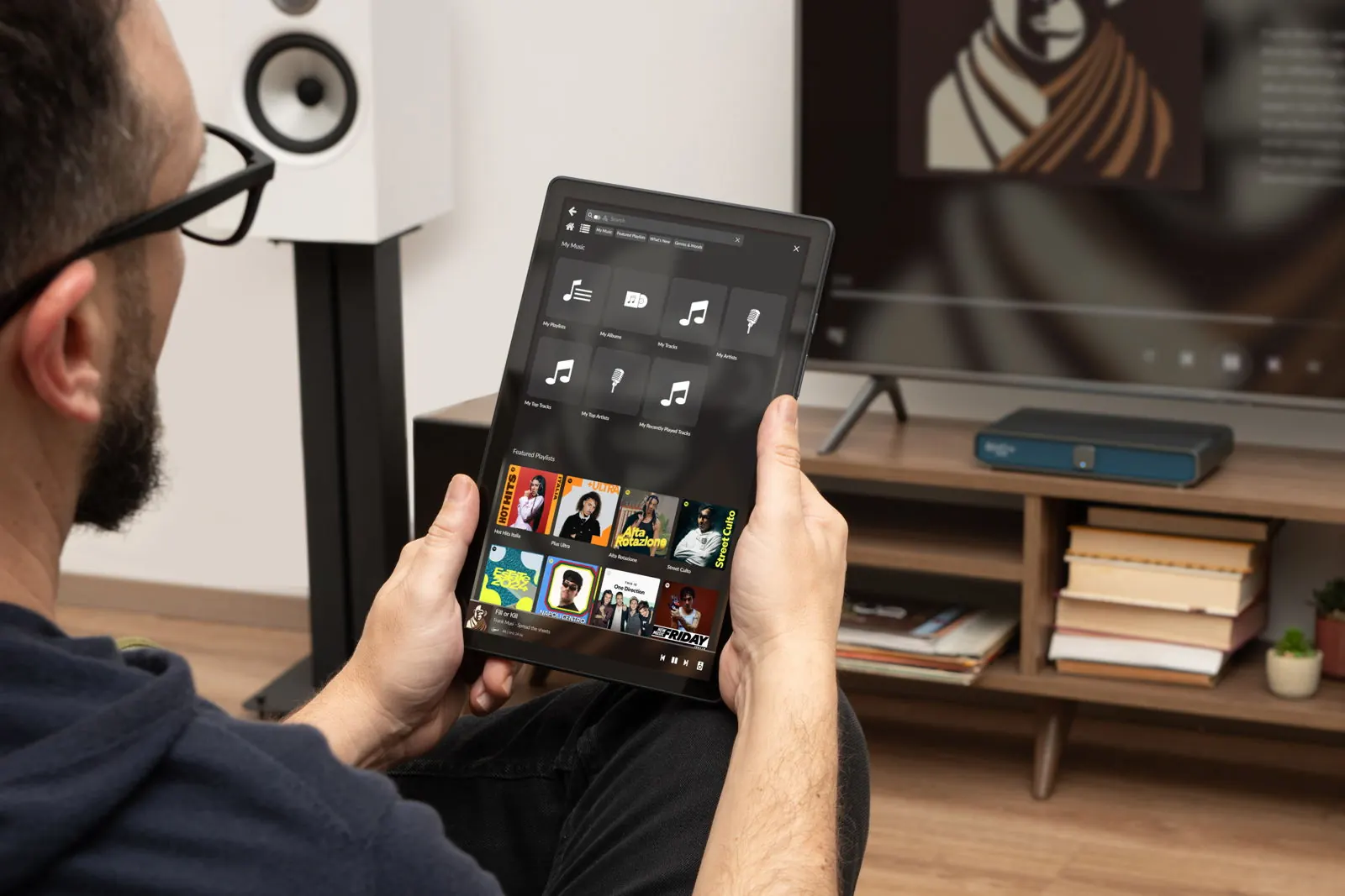 A listener holding a tablet that shows a music library, with Rivo+ on a sideboard under the TV behind him.