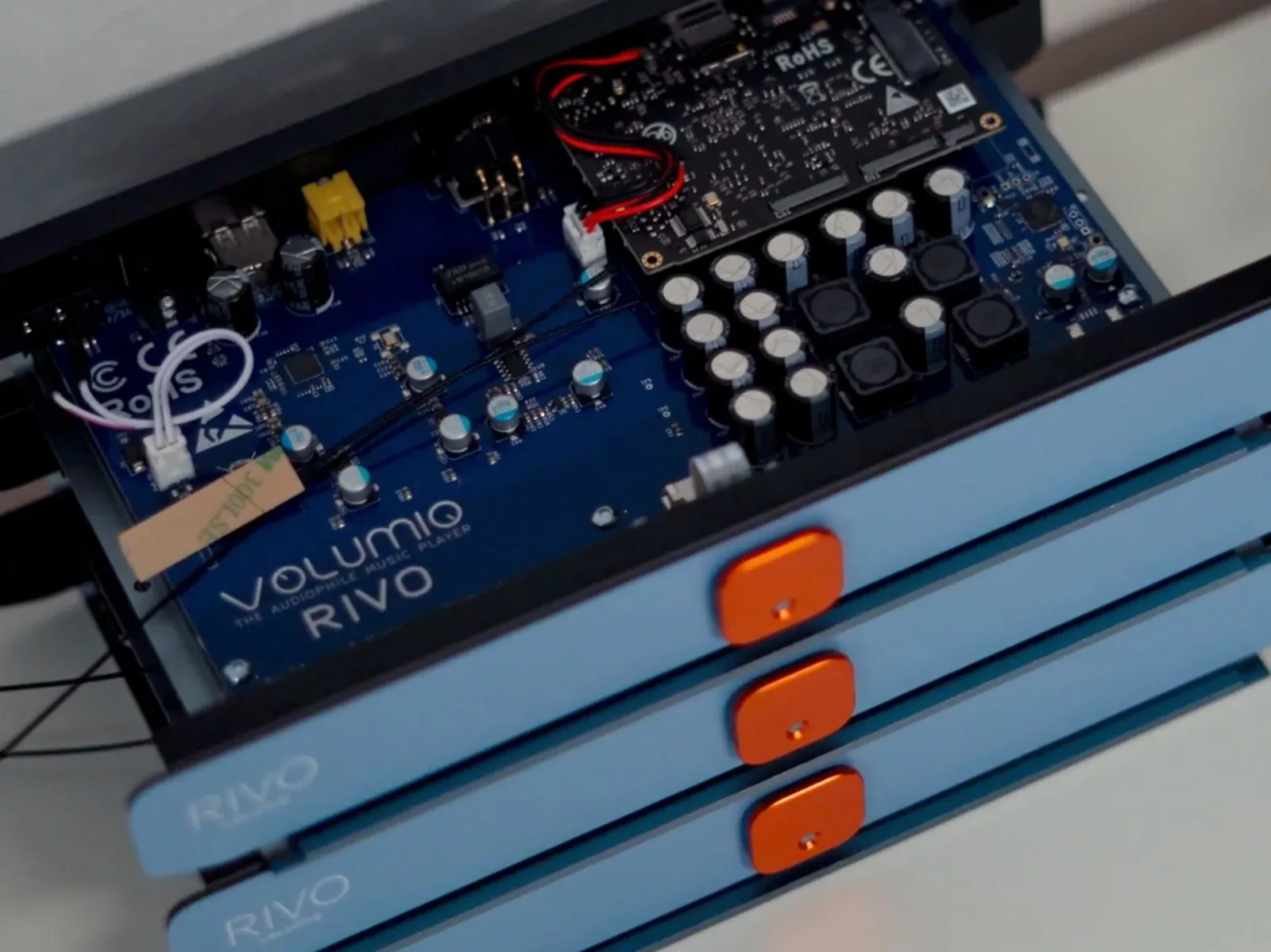 Three Rivo units stacked, with light blue fronts and orange buttons. The top one is open and shows its blue main board, printed with the Volumio Rivo name.